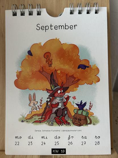 Foto eines Kalenderblatts von Daniela Schreiter aka Fuchskind: Unter einem orange gefärbtem Baum sitzt ein rotes Echsenwesen mit Zaubererhut, in einer Hand hält es einen Apfel, in der anderen einen heißen Tee. Waldtiere beobachten es.