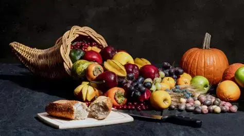 Photo of a cornucopia with an abundance of different fruit flowing from it. A wooden board with bread and a knife is placed in front of it and an orange pumpkin sits in the background.