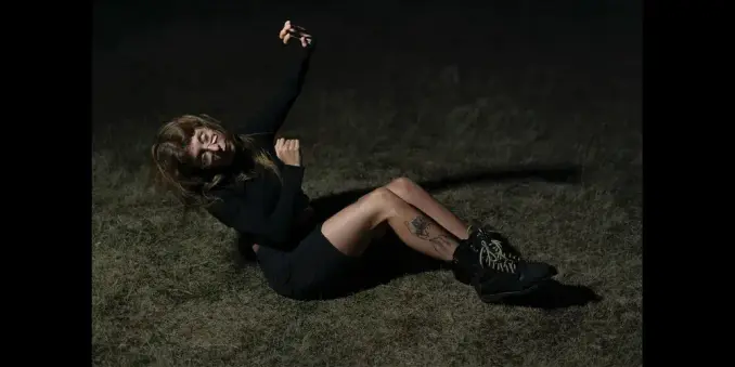 Musician Emma Grace in black lies on dry grass, smiling with eyes closed and arms raised. Custom boots and a leg tattoo are visible in the dark setting.
