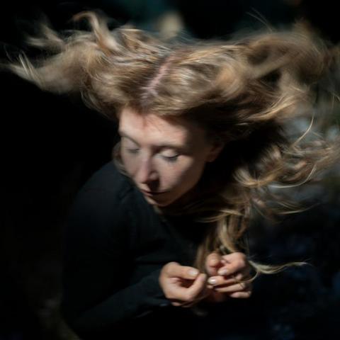 The cover for Emma Grace's Satellites album, showing Grace in black with closed eyes, hair flowing dramatically in motion, set against a dark, blurred background, creating a dreamy mood.

