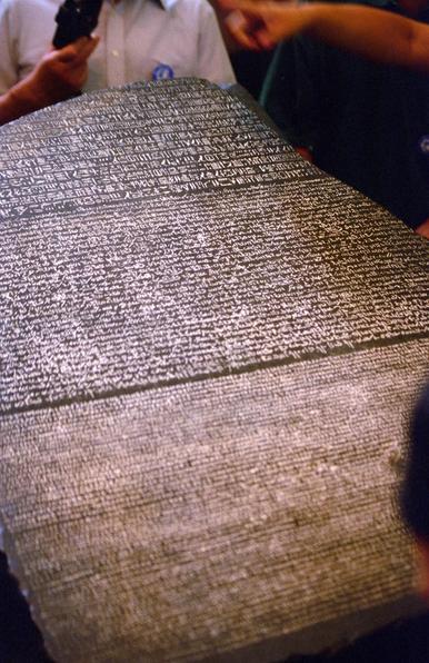 An ancient artifact resembling a stone tablet covered in inscriptions. The text appears to be written in various ancient scripts, with people in the background observing it closely. It is the Rosetta Stone