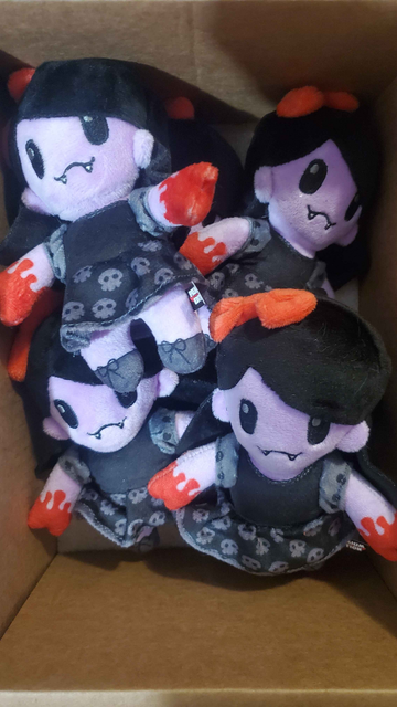 A cardboard box with about a half dozen small plushies of The Clawed Girl from the They Bleed Pixels. She's a purple demon girl with red blood soaked claws in black dress with skulls on it. Her hair is black with a red bow.
