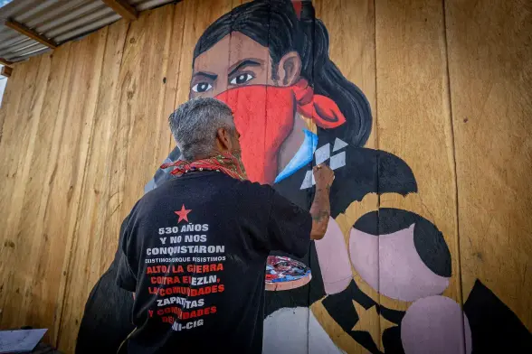 A man with short gray hair and a red bandana around his neck paints a mural on wooden planks. The mural shows a stern-faced Zapatista woman with a red mask, long black hair, and black clothing with geometric patches. His shirt reads in Spanish: “530 años y no nos conquistaron / existimos porque resistimos / Alto a la guerra contra el EZLN, las comunidades zapatistas y comunidades del CNI-CIG.” The wall is raw wood, the ceiling tin, the mood serious and defiant.
