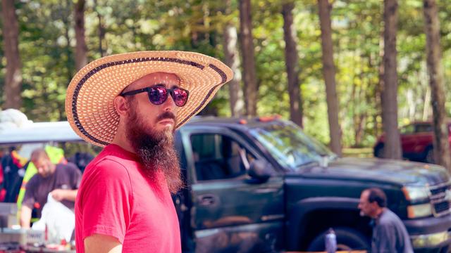 A man with a long beard and wide brimmed straw hat has purple sheen sunglasses and a red shirt walking from the left to the right side with a blue pickup truck,  some other people,  and the stems of tall trees in the background