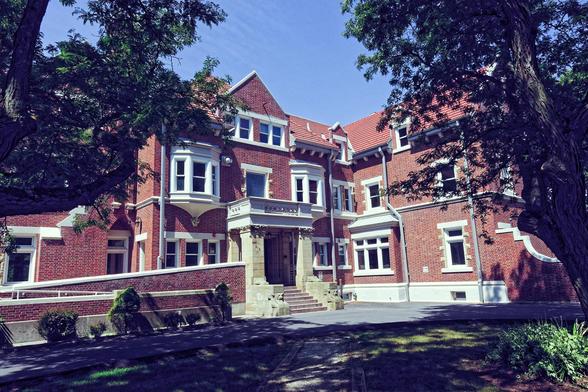 A brick manor of more than three stories with prominent white windows frame by leafed out trees backed by a blue sky