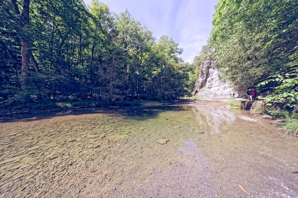 Wide angle shot of a clear pond with a brown rocky bottom with trees on the bank to the left but bounded by an rocky outcropping on the right side with people hiking up a trail cut into the rock.
