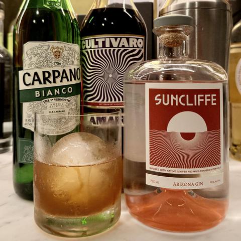 An old fashioned glass filled with a New Age Negroni over a large sphere of ice stands in front of the three ingredients of the cocktail and a metal mixer.