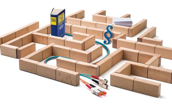 Symbol für die komplexen rechtlichen Rahmenbedingungen in der IT: Zu sehen sind ein Labyrinth aus Holzklötzen mit Kabeln, ein Gesetzbuch, ein Stapel Papier und ein Paragraphenzeichen. 