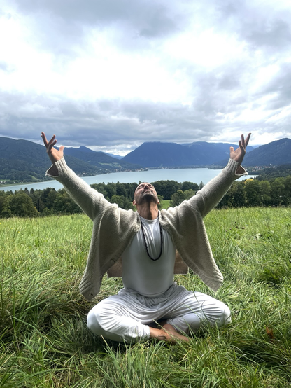 „Ein Mann in weißer Kleidung steht auf einer grünen Wiese vor einem See, breitet die Arme weit zum Himmel aus – Ausdruck von Freude und Freiheit.“