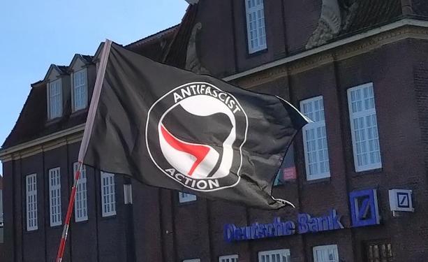 Vor einem Gebäude mit dem Schriftzug Deutsche Bank weht eine schwarze Fahne mit dem rot-schwarz-weißen Symbol der Antifa und den Worten Antifascist Action.
