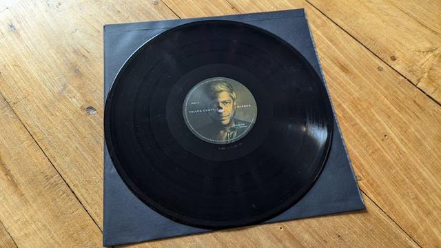 Eine schwarze Schallplatte liegt auf einer hölzernen Unterlage. Das Label zeigt das Porträt eines Mannes und den Text "Trilok Gurtu - Mirror". Es befindet sich neben einer schwarzen Hülle.