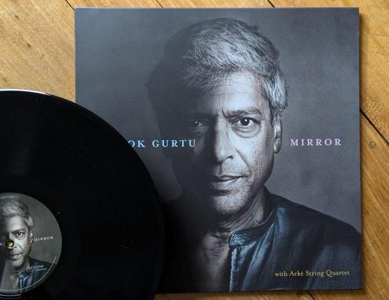 Eine Schallplatte liegt neben ihrem Cover, auf dem das Porträt eines Mannes mit grauem Haar und dunkler Kleidung abgebildet ist. Der Titel des Albums "Mirror" und der Name des Künstlers, "Trilok Gurtu", sind gut sichtbar angebracht.