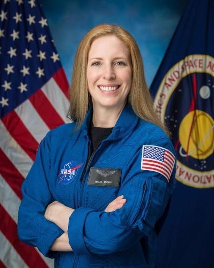 Image Description
Astronaut candidate Imelda Muller, wearing a blue flight suit, looks at the camera with crossed arms. The background is blue with an American flag on the left and a NASA flag on the right. Credit: NASA/Josh Valcarcel