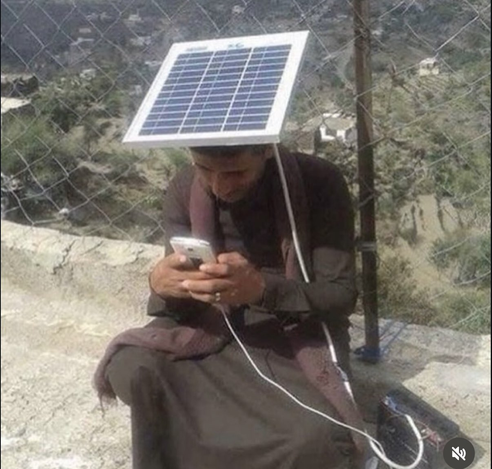 An image of a man wearing a Middle Eastern robe and scarf in rocky terrain wearing a flat square solar panel on top of this head which appears to be charging his smartphone as he interacts with it.