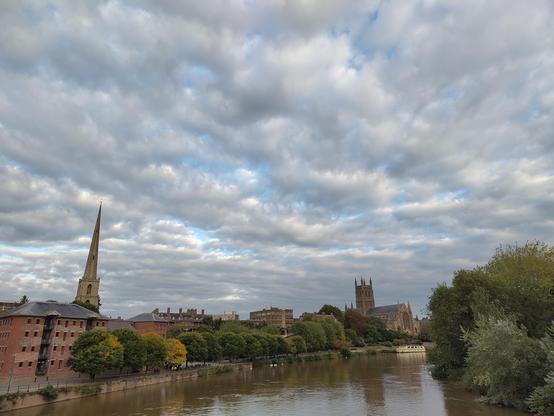 Worcester Cathedral and St Andrew's Spire, locally known as The Glover's Needle, stand tall on the River Severn under a blue Sky with White fluffy Clouds
