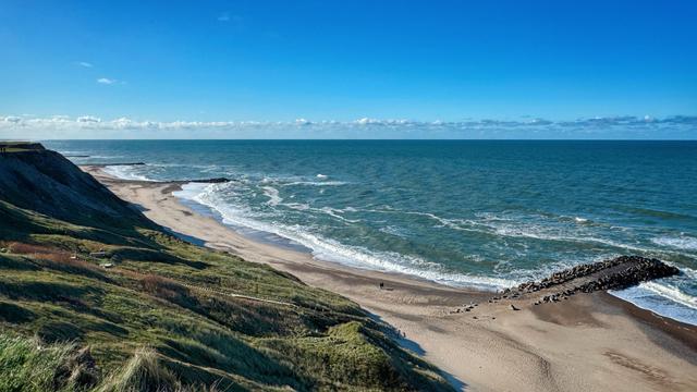 Sicht von begrünter Steilküste auf einen Sandstrand und mehrere Buhnen, an denen sich die Beendung bricht. Der Himmel ist blau, und nur ein kleines Wolkenband ist am Horizont zu sehen. 
