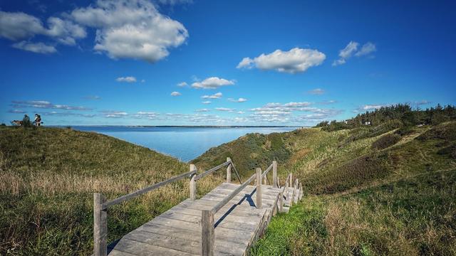 Eine Holztreppe mit Geländer führt an einer Steilküste zwischen hohen Dünen nach unten. Im Hintergrund ist der blau schimmernde Limfjord zwischen den Dünen zu sehen, ebenso ein blauer Himmel mit ein paar kleinen Schäfchenwolken. 