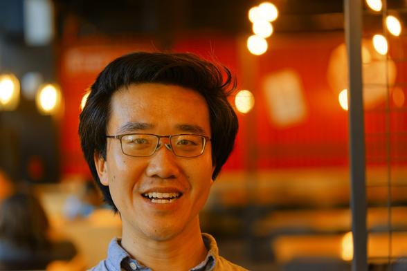 Asian man with close cropped hair has a big smile with a background including bright blurred out lights,  grids and a big red wall