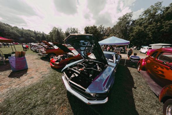 About five cars in a row from the lower center towards the far right with their hoods open more generally surrounded by cars, tents and trees under an unsettled sky.