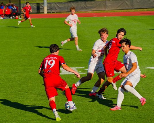 Red Number 19 at the lower center is running to the left with the ball away from a group of 2 white and 1 red player to his left,  while a white player and red player look on from the distance, past the green field can be seen the red surface of the track and a fence and just a bit of the player bopx