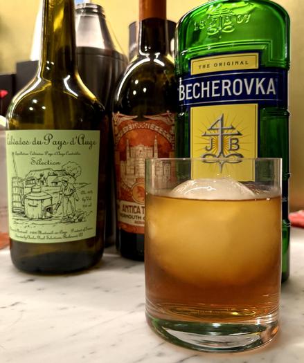 An old fashioned glass filled with a Calcherovka Negroni over a large sphere of ice stands in front of the three ingredients of the cocktail and a metal mixer.