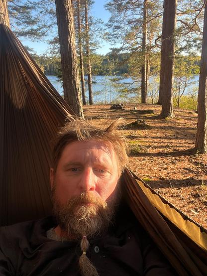 Selfie of yours truly in their hammock next to a lake between trees. Ground is covered in golden brown needles and leaves. 🍂 