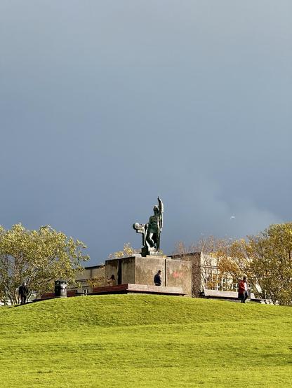 Arnarhóll, a nice green space in the city center, is bathed in sunlight with dark skies behind. The statue of Ingólfur Arnarson can be seen at the top of the hill