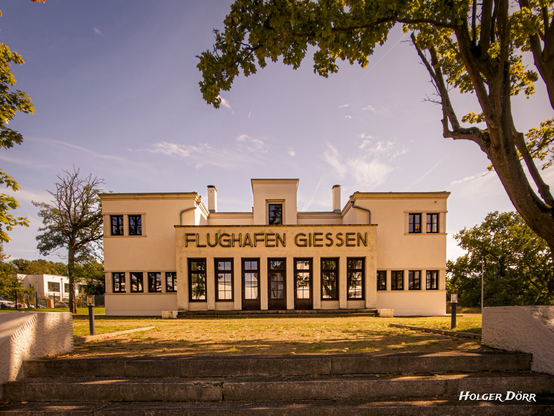 Sommerliches Foto des Alten Flughafens in Gießen. Das historische Gebäude wirkt in der warmen Sonne besonders eindrucksvoll, klare Linien und helle Farben verleihen der Szene eine stille Würde. Ein Ort, der Vergangenheit und Gegenwart verbindet.