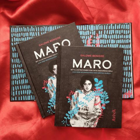 Drei Exemplare von "Maro" liegen auf einem roten Tuch: eines aufgeschlagen, so dass der Vorsatz zu sehen ist, darauf zwei geschlossene. Das Cover zeigt eine junge Frau vor schwarzem Hintergrund.