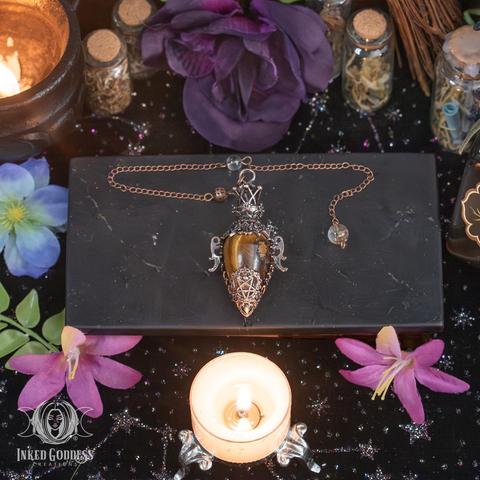An example of the "Tiger's Eye Witchy Pendulum" product is laid out on a black stone surface. It features a teardrop-shaped tiger's eye gemstone suspended from a copper chain and mounted in silver and copper metal filigree. Presented by Inked Goddess Creations.