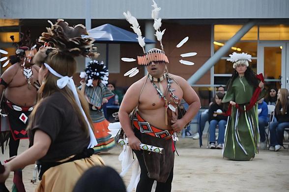 A man wearing traditional regalia with feathered headdress, beadwork, and face adornments performs in a tribal dance, surrounded by other dancers and onlookers.