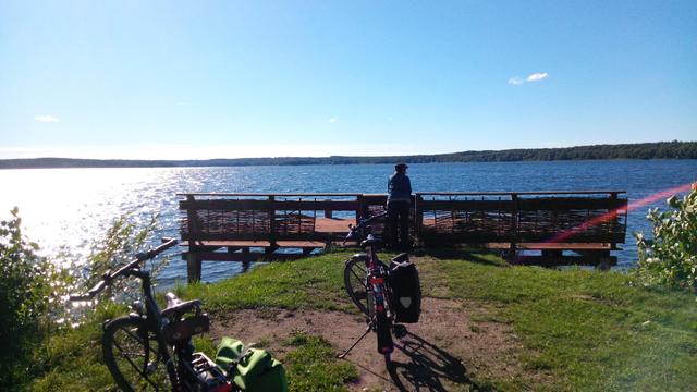 2 Fahrräder, 1 Person am Anlegergeländer, dahinter blaues Wasser, links ins silber übergehend durch Sonnenreflektion