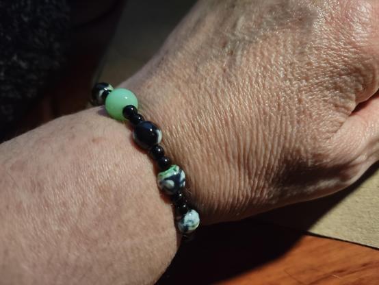 picture of bracelet on a wrist. showing partial arm and partial hand. bracelet is beaded. small round black beads in between larger round black and green swirled beads. a single round larger bead sits at top of wrist. it is a glow in the dark shade of green and made with uranium dioxide. not seen: a small uranium dioxide green heart bead on bottom of wrist.