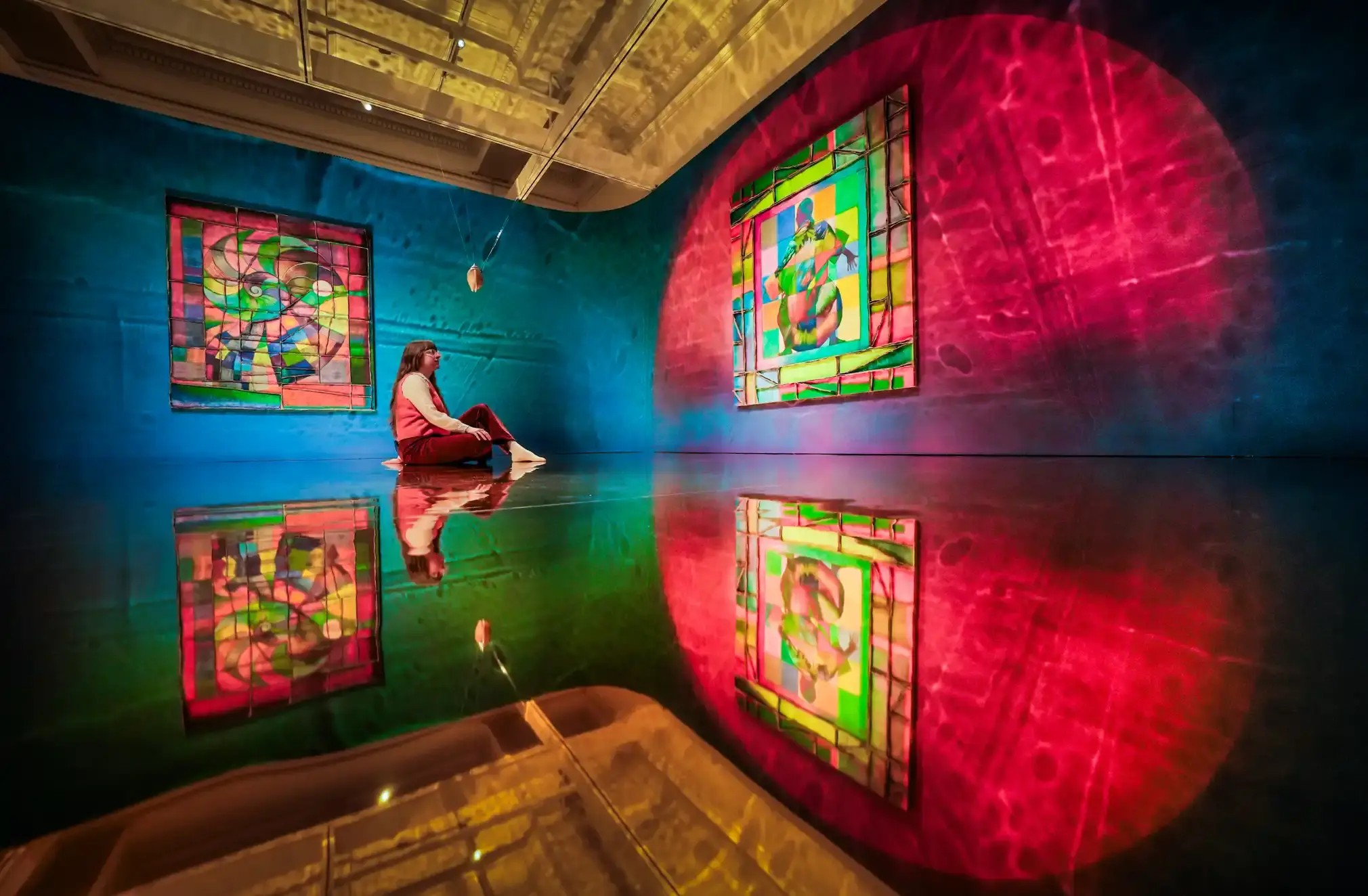 A woman sits in a mirrored gallery in front of an artwork.