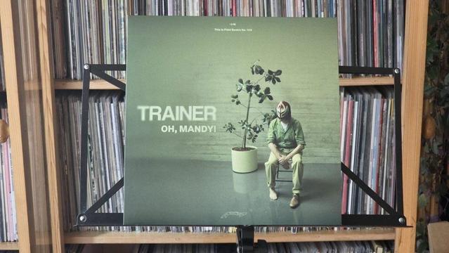 Ein Schallplattencover mit dem Titel "TRAINER - OH, MANDY!" zeigt eine Figur, die auf einem Stuhl sitzt, eine Maske und Freizeitkleidung trägt und neben einer Topfpflanze steht. Die Szene ist vor einem strukturierten Hintergrund mit Regalen voller Schallplatten zu sehen.