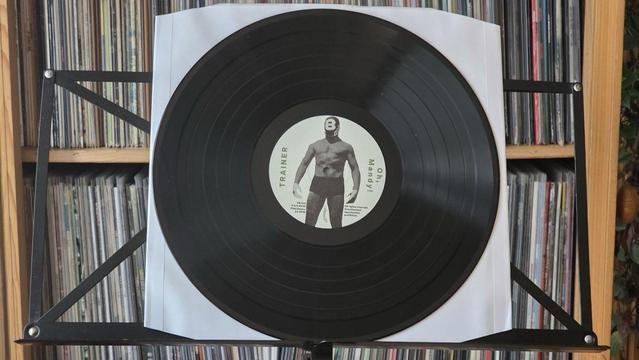 Auf einem Ständer steht eine Schallplatte mit einem Etikett, auf dem eine Person mit einer Wrestling-Maske abgebildet ist und der Text "TRAINER" und "Oh, Mandy!" in einem kreisförmigen Muster steht. Im Hintergrund befinden sich Platten-Regale.