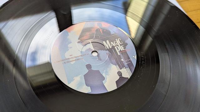 Nahaufnahme einer Schallplatte mit dem Etikett des Albums "Magic Pie". Das Etikett enthält eine künstlerische Grafik mit zwei Silhouetten und Text, der den Titel und den Liedtext angibt. Die Schallplatte liegt auf einer Holzfläche mit natürlichem Licht.