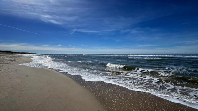 Nordseestrand an einem sonnigen Tag. Die Brandung fällt milde aus, am Himmel sind nur ein paar Schleierwolken zu sehen. 