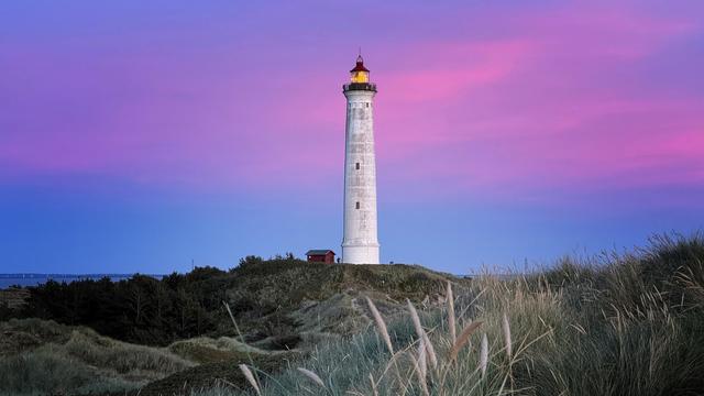 Weißer Leuchtturm mit Leuchtfeuer ragt aus den Dünen hervor, im Hintergrund schimmert der Himmel nach Sonnenuntergang Blau, Rosa und Lila. 