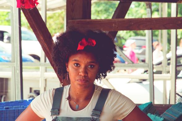 African-American woman with curly hair shaped into a big sphereoid with a red bow smack in the middle.  She's wearing and we can see the straps and very top of the front of denim overalls,  behind are some wood planks of the farmer's market structure and behind it blow out cars and trees since it is much brighter outside.