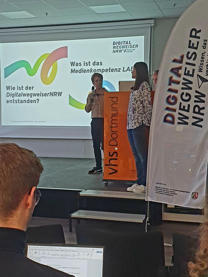 Das Bild zeigt eine Veranstaltung in der vhs Dortmund, bei der eine Präsentation zum Thema "Medienkompetenz Lab" stattfindet. Auf einem großen Bildschirm hinter der Bühne ist der Titel "Was ist das Medienkompetenz Lab?" zu sehen, zusammen mit der Frage "Wie ist der DigitalwegweiserNRW entstanden?" und dem Logo des Digitalwegweisers NRW. Zwei Personen stehen auf der Bühne, eine Frau hält ein Mikrofon und spricht, während die andere Person neben ihr steht. Ein Banner mit dem Text "DIGITAL WEGWEIS…