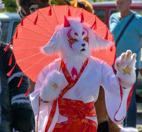 Ein Kitsune Fursuiter mit roten Sonnenschirm in einer Fursuit Parade