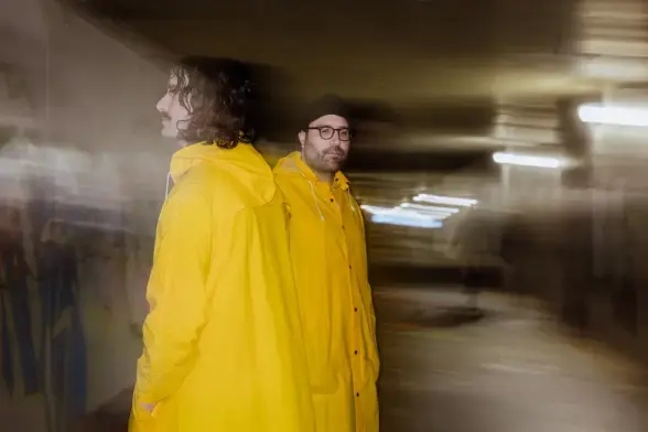 Daniel Lyon and Daniel “Connor” Birch aka Packaging, in yellow jackets stand in an underpass with blurry lights. One faces away, the other looks forward. Mood is mysterious.
