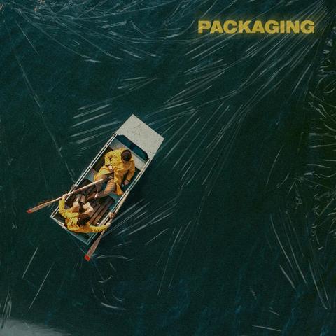 The cover for Packaging's self titled album, showing the two band members in yellow raincoats rowing a small boat on a dark, cling film covered body water. The word "PACKAGING" is visible in the top right corner.
