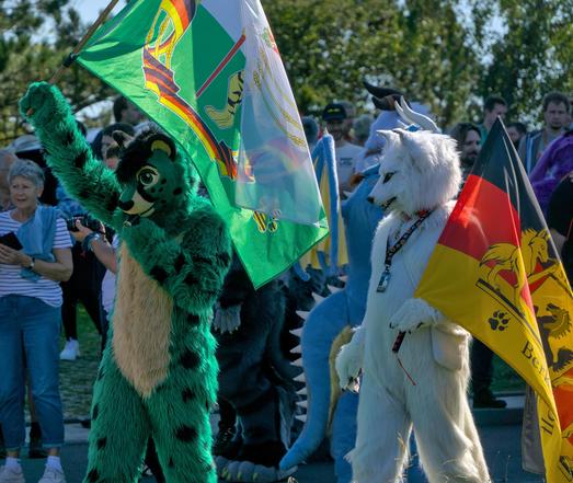Ein grüner Katzen Fursuiter und ein weißer Wolf Fursuiter schwingen thematischen Flaggen
