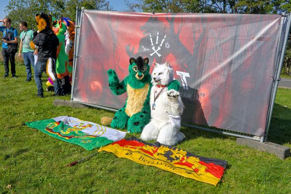 Grüner Katzenfursuiter mit Flagge und weißer Wolf Fursuiter mit Flagge sitzen vor dem EAST 13 Logo