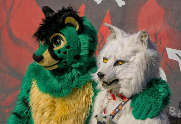 Grüner Katzenfursuiter und weißer Wolf Fursuiter sitzen vor dem EAST 13 Logo.
Der Katzenfursuiter hat seinen arm um den Wolf Fursuiter gelegt