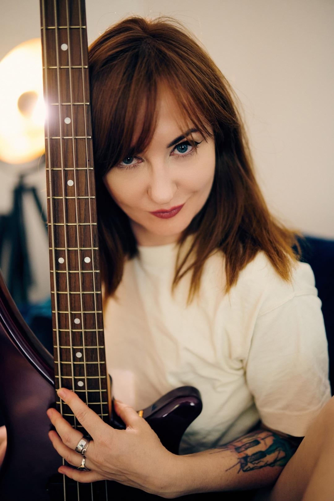 Close-up of Kat de Ville holding a bass guitar horizontally, the camera focusing along the fretboard towards her confident expression in the background. Close-up of Kat de Ville holding a bass guitar horizontally, the camera focusing along the fretboard towards her confident expression in the background.