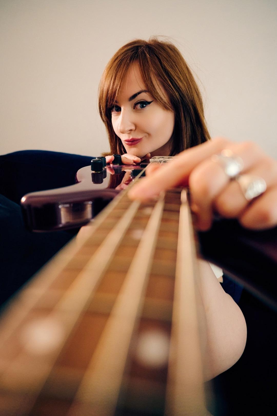Kat de Ville sits indoors, hugging the neck of the bass guitar close to her face, a soft light in the background casting a warm glow. Kat de Ville sits indoors, hugging the neck of the bass guitar close to her face, a soft light in the background casting a warm glow.