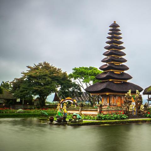 Frist part of panorama split in two: Eleven-tiered Meru towers of Pura Ulun Danu Beratan rising from the calm waters of Lake Beratan, shrouded in mist with forested hills and a cloudy sky behind.